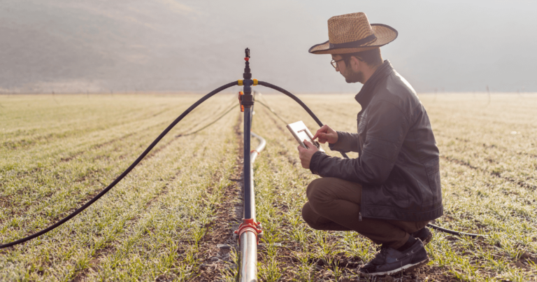 Agricultor utilizando tablet para monitorar sistema de irrigação em lavoura, representando práticas de agricultura moderna e uso de tecnologia no campo.