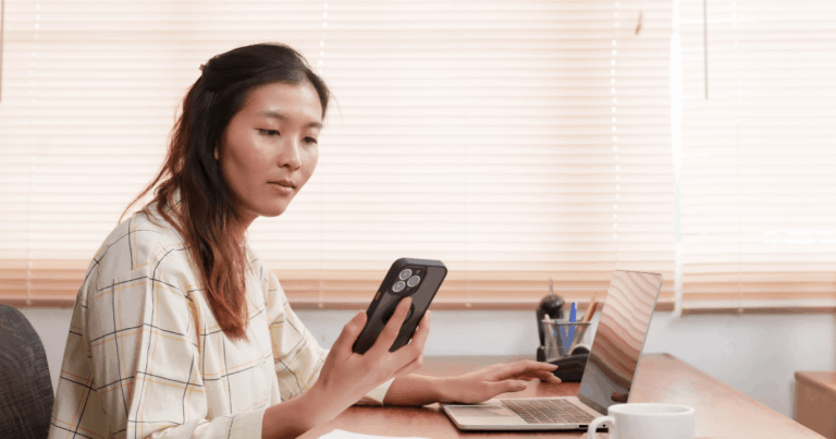 Mulher concentrada usando celular e notebook em ambiente de trabalho moderno, ilustrando o uso prático do DDA (Débito Direto Autorizado) para gestão financeira eficiente