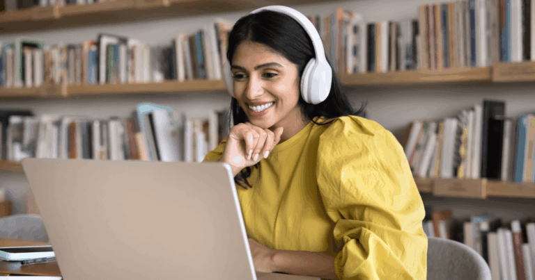 Mulher sorridente usando fones de ouvido e assistindo a um curso online em seu notebook, representando o conceito de e-learning em ambiente de estudo com estante de livros ao fundo.