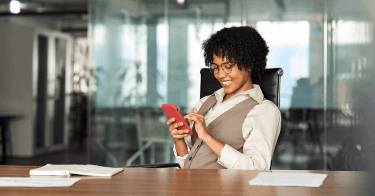 Profissional sorrindo enquanto utiliza o celular em um ambiente de escritório, representando a interação com mensagens automáticas no WhatsApp.