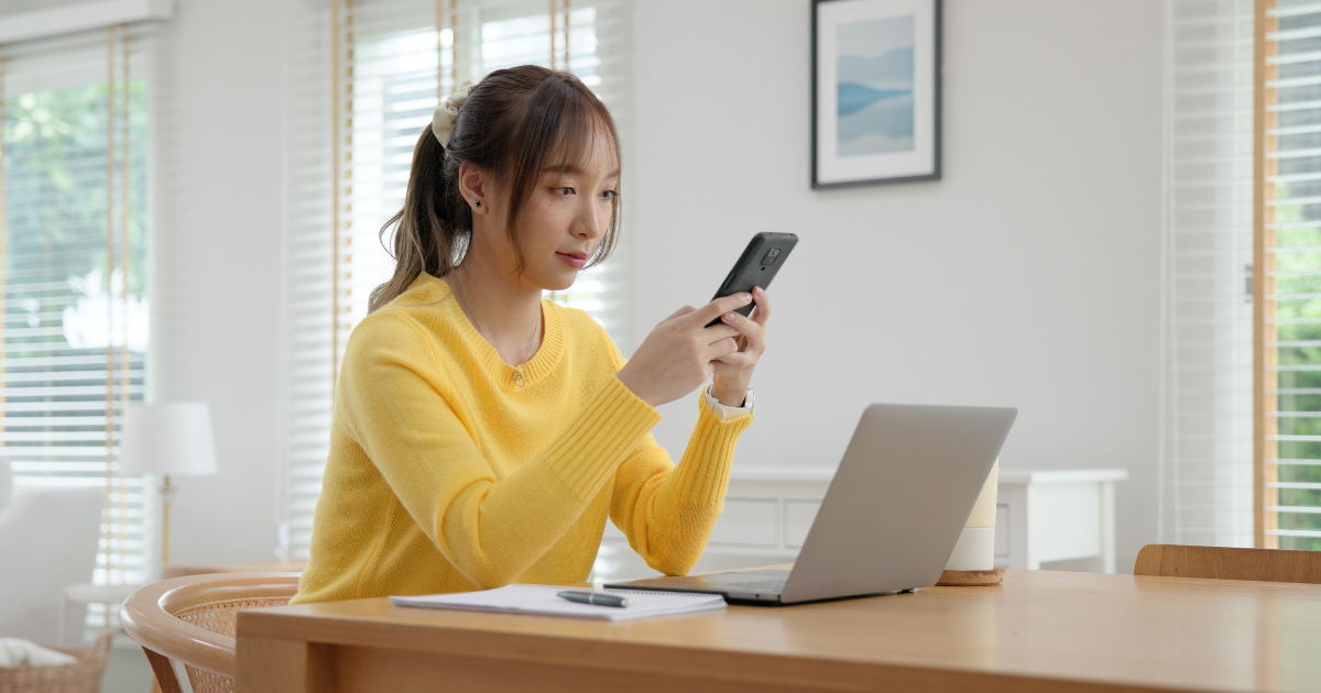 Mulher usando chatbot no WhatsApp pelo celular, em ambiente de home office com notebook e bloco de anotações sobre a mesa.