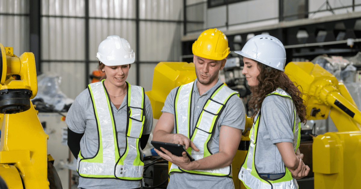 Trabalhadores com capacetes e coletes de segurança analisando dados em tablet dentro de indústria, representando práticas de segurança no trabalho conforme a NR-9.