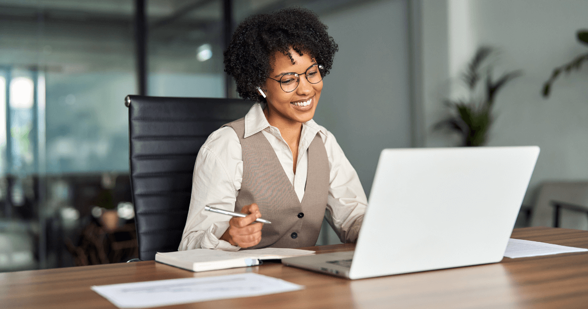 Profissional sorrindo durante reunião one a one online, com notebook, anotações e fone de ouvido, representando a prática de 1:1 nas empresas.